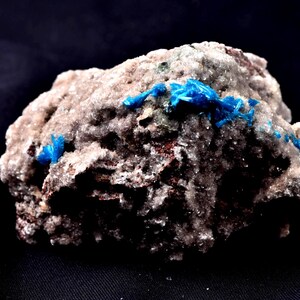 May include: A close-up of a mineral specimen. The image shows a rough, grey and brown rock formation with bright blue, crystalline structures embedded within. The contrast is striking against the black background.