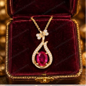 May include: A gold pendant necklace featuring a central oval-cut vibrant pink gemstone surrounded by a curved frame of small clear stones, with a small floral-shaped accent at the top, resting inside a deep red velvet jewelry box.