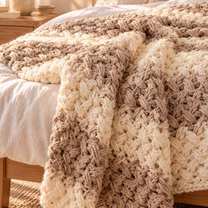 May include: A chunky knit throw blanket featuring a thick, braided cable pattern in alternating stripes of cream and light brown yarn. The blanket is draped over the edge of a bed, showcasing the textured, looped construction of the fabric.