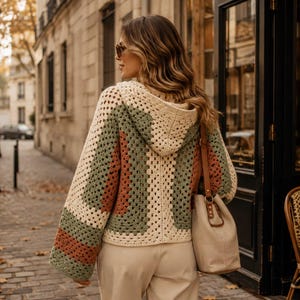 Op de afbeelding: Een gehaakte trui met capuchon en een granny square patroon in crème, saliegroen en terracotta. Het kledingstuk heeft lange, wijde mouwen en een capuchon, gecombineerd met een beige schoudertas en een lichte broek, gezien vanaf de achterkant.