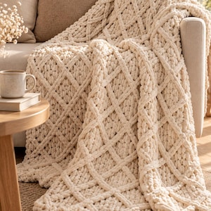 Può includere: Una coperta in maglia grossa color crema con un motivo a rombi in rilievo. La coperta è realizzata in filato spesso e morbido ed è drappeggiata sul bracciolo di un divano, mettendo in risalto la sua struttura a trecce testurizzata.