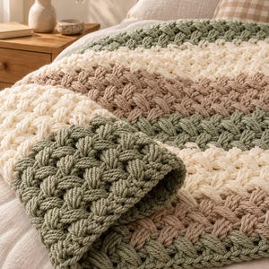 May include: A chunky knit throw blanket featuring a thick, braided cable pattern with horizontal stripes in alternating shades of sage green, cream, and light tan. The heavy yarn texture creates a raised, woven appearance across the surface.