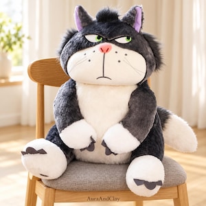 May include: A plush cat toy with dark gray fur, a white belly, and white paws. The cat has a grumpy facial expression with green eyes, a pink nose, and black whiskers. The text AuraAndClay is printed on the wooden chair seat below the toy.