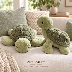 Puede incluir: Dos tortugas de peluche color verde salvia con pelaje suave y texturizado y patrones de caparazón detallados. Una tortuga está acostada y la otra sentada. El texto AuraAndClay es visible en la parte inferior con la frase Styles shown.