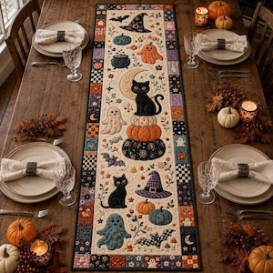 Puede incluir: Un camino de mesa rectangular de tela con un borde de patchwork y un panel central con motivos de Halloween que incluyen gatos negros, fantasmas, calabazas, sombreros de bruja, murciélagos y una luna creciente sobre un fondo crema.