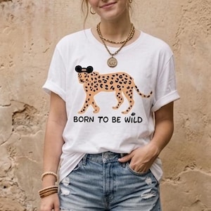 May include: White t-shirt with a leopard graphic wearing sunglasses and the text "BORN TO BE WILD". The shirt is paired with denim shorts. The model is wearing gold jewelry and bracelets.