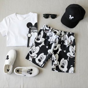 A casual outfit consisting of a white t-shirt with a Mickey Mouse design, black and white shorts, a black baseball cap, a watch, and a pair of white sneakers with Mickey Mouse designs.