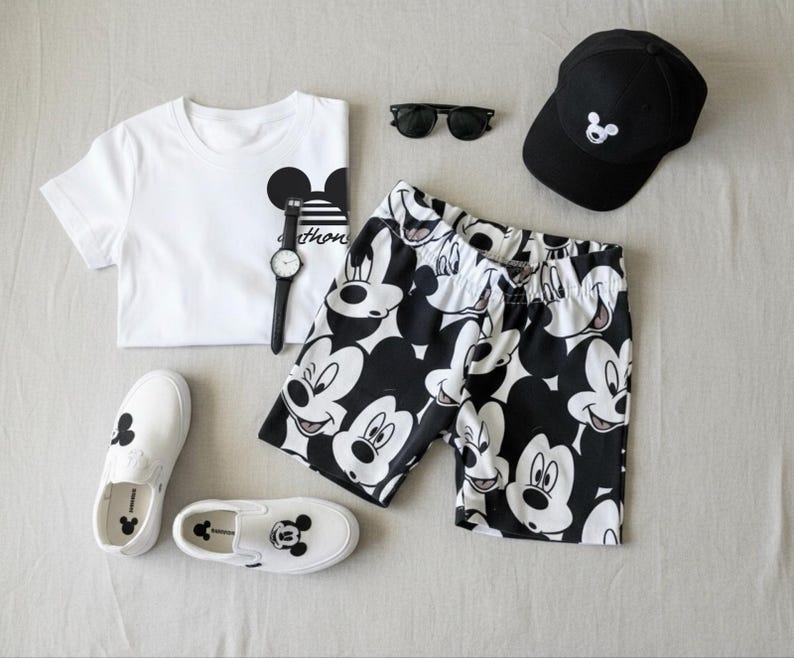 A casual outfit consisting of a white t-shirt with a Mickey Mouse design, black and white shorts, a black baseball cap, a watch, and a pair of white sneakers with Mickey Mouse designs.