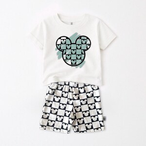 A white t-shirt with a graphic of a Mickey Mouse design and a pair of shorts with a repeating pattern of black and white Mickey Mouse icons.