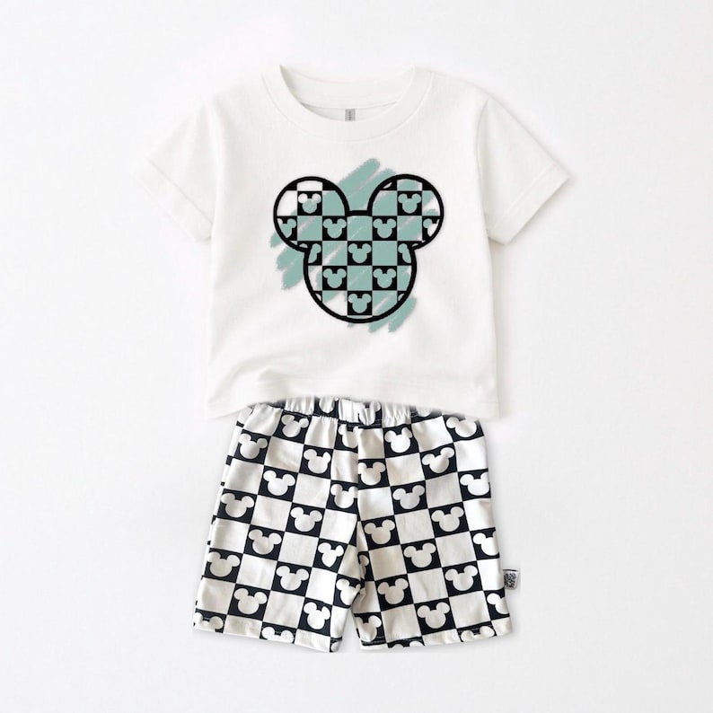 A white t-shirt with a graphic of a Mickey Mouse design and a pair of shorts with a repeating pattern of black and white Mickey Mouse icons.