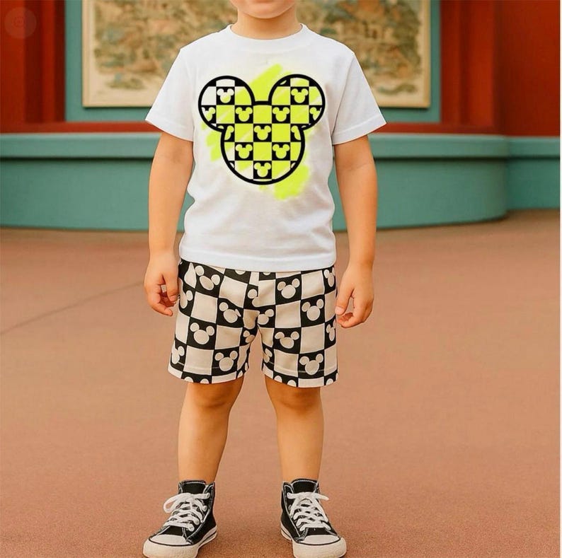 A young boy wearing a white t-shirt with a Mickey Mouse design, black and white shorts, and black and white sneakers.