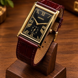 Op de afbeelding: Een rechthoekig Seiko-polshorloge met een goudkleurige kast en een zwarte wijzerplaat met gouden Romeinse cijfers. Het horloge heeft een donkerbordeauxrode lederen band met een textuur en een kleine sub-wijzerplaat voor de seconden.