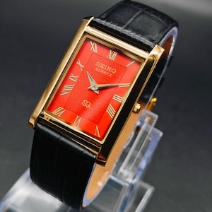 May include: A rectangular Seiko Quartz wristwatch featuring a vibrant red dial with a wavy guilloche pattern, gold-tone Roman numeral hour markers, and gold-tone hands. The watch has a polished gold-tone case and a black leather strap.