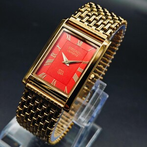 May include: A rectangular gold-tone wristwatch featuring a vibrant red dial with a textured wave pattern. The watch face displays gold Roman numeral hour markers and gold hands. It includes a matching gold-tone metal link bracelet strap.