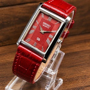 Könnte beinhalten: Eine rechteckige Seiko Quartz Armbanduhr mit einem leuchtend roten Zifferblatt mit strukturiertem Wellenmuster, silberfarbenen römischen Ziffern als Stundenmarkierungen und silbernen Zeigern. Die Uhr hat ein glänzendes rotes Lederarmband.