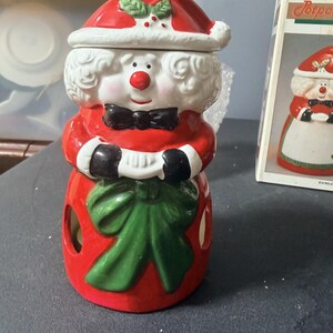 May include: A ceramic Mrs. Claus candle warmer featuring a red Santa hat with holly, white curly hair, a red nose, and a black bow tie. The figure wears a red dress with a large green bow at the waist and has cut-out openings on the sides.
