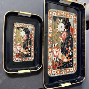 May include: Two rectangular black serving trays with rounded corners and gold-toned rims. Each tray features a central decorative panel with a peacock, white and orange floral motifs, and a border of small orange and white flowers on a gold background.