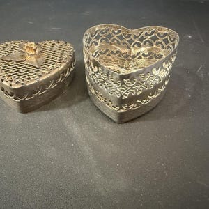 May include: Two heart-shaped silver-toned metal containers. One container is a lidded box with a mesh top and a central rose ornament. The other is an open-work basket with a decorative filigree pattern along the sides and a heart-shaped rim.