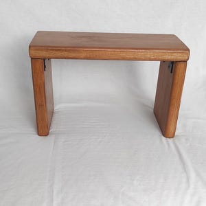 Puede incluir: Un banco rectangular de madera con un acabado liso de color marrón medio. El banco cuenta con una superficie superior plana sostenida por dos paneles laterales verticales, con pequeñas bisagras de metal oscuro en las esquinas interiores.