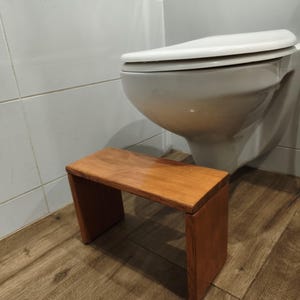 Peut inclure: Un marchepied rectangulaire en bois avec une finition lisse brun moyen. Le marchepied présente une surface supérieure plate soutenue par deux panneaux latéraux verticaux, créant une structure simple à cadre ouvert posée au sol.
