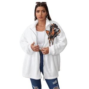 May include: A white zip-up fleece jacket featuring a high collar and a graphic print on the upper left chest. The graphic depicts a stylized cowboy figure in black and orange tones riding a horse. The jacket has long sleeves and a soft texture.