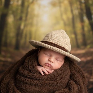 May include: A sleeping baby wrapped in a brown blanket, wearing a beige crochet hat with a brown band. The hat has a wide brim and a small buckle detail. The background is a blurred forest scene.