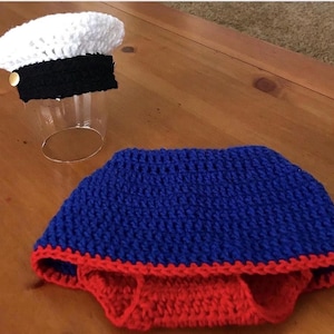 May include: Crocheted baby sailor outfit, featuring a white and black sailor hat with a gold button, and a blue and red diaper cover. The handmade set is likely for a newborn photo prop or costume.
