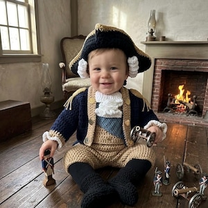 May include: A baby wearing a crocheted colonial costume, including a tricorn hat, jacket, and pants. The navy blue jacket has gold buttons and trim. The baby is holding a toy cannon and is surrounded by toy soldiers and cannons.