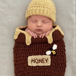 May include: A newborn baby wearing a crocheted honey pot costume with a matching hat. The brown costume has a beige honey drip design and a "HONEY" label. A small bee applique is on the front.
