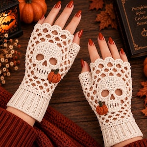 May include: A pair of off-white fingerless gloves featuring a crocheted skull pattern on the back of the hand and a small, textured orange pumpkin accent. The gloves have a ribbed cuff at the wrist and a scalloped edge along the finger openings.