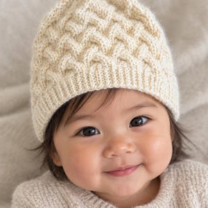 Pode incluir: Um gorro de malha na cor creme com um padrão de tranças texturizado e uma borda canelada. O gorro é feito de fio e apresenta um design geométrico repetitivo na coroa, proporcionando um ajuste justo com uma construção clássica.
