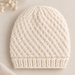 Pode incluir: Um gorro de malha na cor creme com um padrão de pontos diagonais texturizados no corpo principal e uma borda de malha canelada na base. O gorro é feito de fio grosso e apresenta uma cor branca suja consistente e uniforme.