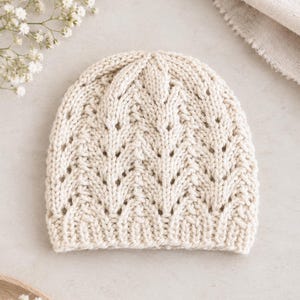 May include: A cream-colored knit beanie featuring a repeating vertical chevron pattern with openwork eyelet details. The hat has a ribbed edge at the bottom and a rounded crown, constructed from a thick yarn with a visible textured stitch pattern.