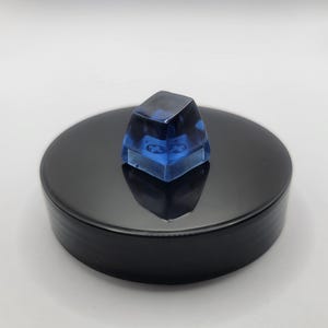 May include: A translucent blue resin mechanical keyboard keycap with a smooth, glossy finish and a square top profile. The keycap features a standard cross-shaped stem mount on the underside, visible through the clear blue material.