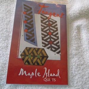 May include: A red booklet with the title "Tangent" and the text "Maple Island Quilts". The booklet features a geometric pattern with black, white, red, and blue colors.