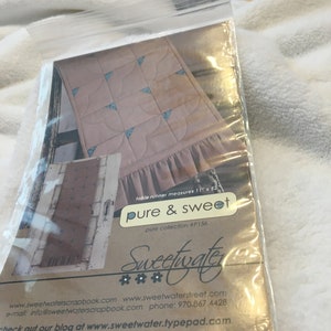 May include: A brown paper package with a plastic window showing a pink and blue quilt pattern. The package is labeled "pure & sweet" and "Sweetwater" with contact information and a website address.