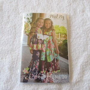 May include: Pink Fig Patterns apron top and dress sewing pattern No. 20. Two girls are wearing the finished garments. The girls are wearing floral print dresses with ruffles. The dress on the left is a dark purple and pink floral print. The dress on the right is a light blue and pink floral print.