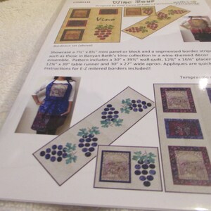 May include: A quilting pattern for a wine-themed decor ensemble. The pattern includes a 30-inch by 39-inch wall quilt, 12-inch by 16-inch placemats, a 12-inch by 39-inch table runner, and a 30-inch by 27-inch apron. The pattern features a design of grapes and the word "Vino".