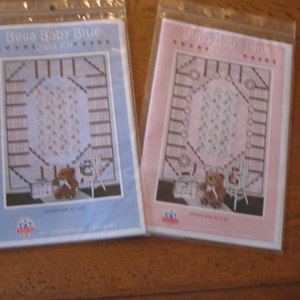 May include: Two quilt kits, one in blue and one in pink, with the text "Bella Baby Blue Quilt Kit" and "Bella Baby Pink Quilt Kit". Each kit features a brown teddy bear, a white cot, and a white quilt with a brown and white pattern.