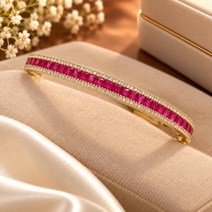 May include: A gold-toned hinged bangle bracelet featuring a central row of square-cut vibrant pink stones flanked by two delicate rows of small, clear pavé-set stones. The piece has a polished metallic finish and a secure clasp mechanism.