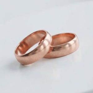 Könnte beinhalten: Zwei Ringe aus Roségold mit einer glatten, polierten Oberfläche. Die Ringe haben ein schlichtes, abgerundetes Profil mit gleichbleibender Breite über den gesamten Umfang und zeigen einen warmen Metallton sowie eine reflektierende Außenseite.