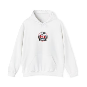 Peut inclure: Un sweat à capuche blanc avec un motif circulaire sur la poitrine. Le graphique montre un drapeau canadien, des montagnes et des pins, avec le texte "I AM CANADIAN" en lettres noires arquées autour du design. Le sweat a des manches longues.
