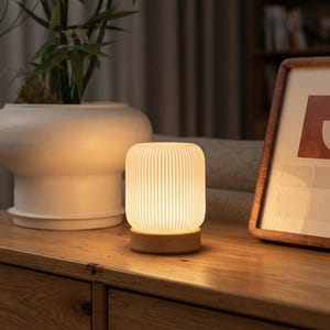 May include: A cylindrical table lamp with a white, vertically ribbed shade and a circular light-colored wooden base. The lamp emits a warm, soft glow, illuminating the surface it rests upon with a gentle light, creating a cozy and inviting atmosphere.