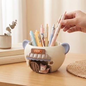 May include: A cream-coloured ceramic pen holder shaped like a small cafe building with a light blue roof and ears. The front features a brown sign with the word CAFE, a small arched door, and a tiny fox figure. It holds several assorted pens.
