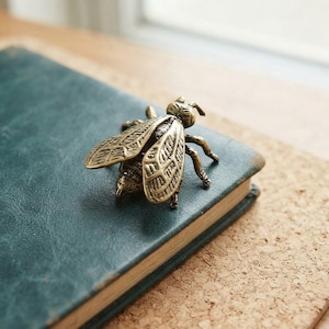 Puede incluir: Una figura de abeja de metal en tono latón con alas grabadas detalladas y un cuerpo segmentado. La escultura del insecto tiene un acabado antiguo y desgastado con una pátina oscura visible en las grietas, mostrando una forma realista.