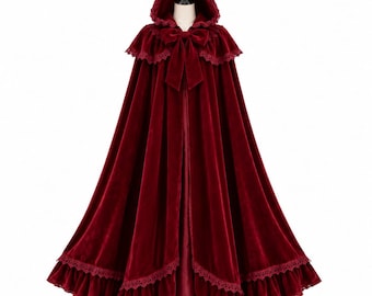 Pode incluir: Uma capa de veludo vermelho profundo até ao chão com capuz pontiagudo, um grande laço de veludo no decote e uma bainha com folhos decorada com detalhes em renda. A peça é exibida num manequim, realçando a sua silhueta ampla e fluida.