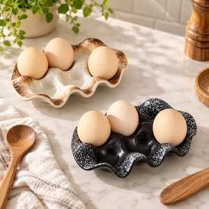 May include: Two ceramic egg holders with wavy edges, one in a cream color with brown rim detailing and the other in a black speckled finish, each holding three light brown eggs in molded indentations for secure storage.