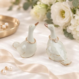 May include: Two pale green ceramic duck-shaped ring holders with brown-tipped beaks. One holder features a plain gold-tone band around its neck, while the other holds a gold-tone ring with a small, clear, oval-shaped stone setting.