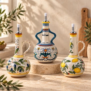 Può includere: Tre oliere in ceramica con beccucci in metallo. L'oliera a sinistra presenta olive blu, quella centrale ha motivi floreali blu e gialli con la scritta OLIO, e quella a destra mostra limoni gialli e foglie verdi su uno sfondo color crema.