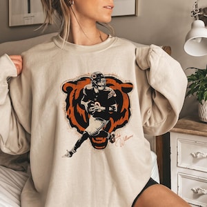 May include: A beige crewneck sweatshirt featuring a graphic of a football player in a dark uniform running with a ball. Behind the player is a large orange and black bear head silhouette. The text Caleb Williams is written in cursive script.